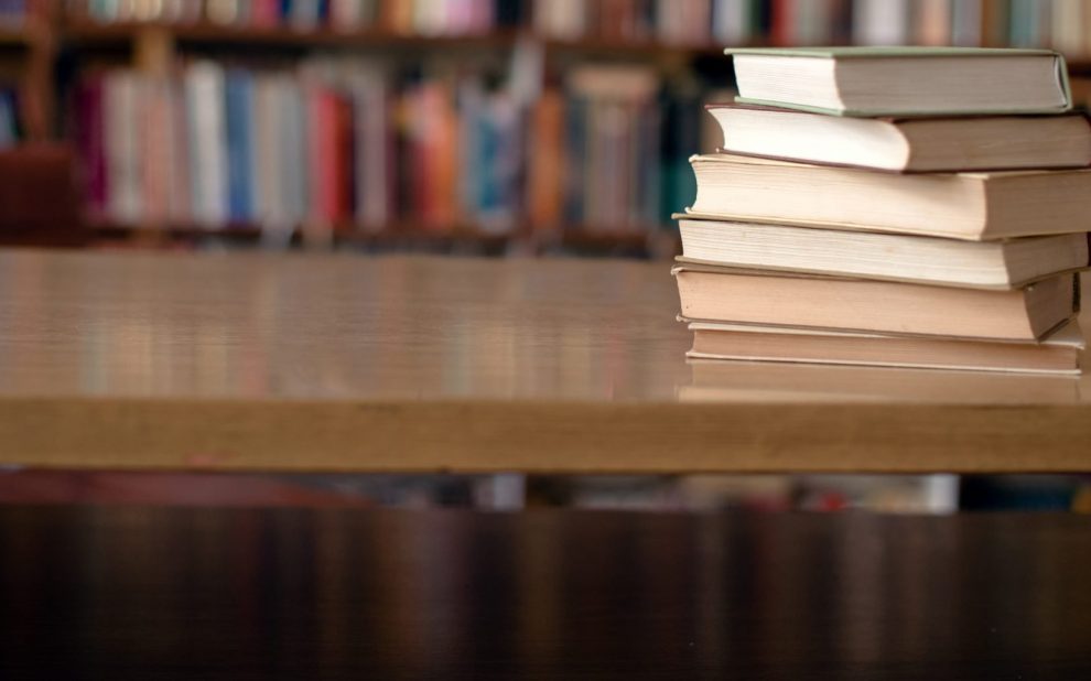 A stack of books on a table in front of a filled bookshelf